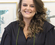 Imagem da nova defensora pública, Gabriela Vizel Gomes. Ela olha para frente, sorrindo. Está com os cabelos soltos, que são cacheados, um pouco abaixo do ombro. Ela usa uma beca preta, com um cordão verde.