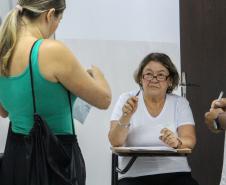 Imagem que mostra uma candidata entrando na sala de aplicação de prova, ela, que está em pé, apresenta os documentos a uma fiscal, que está sentada.