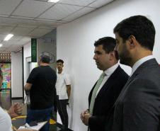 Imagem que mostra dois defensores públicos representantes da comissão organizadora do concurso conversando com uma fiscal no corredor. Atrás deles, candidatos transitam em direção às salas.