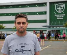 Imagem que mostra o candidato João Carlos de Carvalho em frente ao local de prova, um dos prédios da Universidade Tuiuti, em Curitiba. Ele veste uma camiseta cinza e olha para a frente.