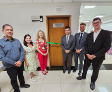 Imagem que mostra a inauguração da sala da DPE-PR em Cruzeiro do Oeste. Estão presentes a juíza diretora do Fórum, a promotora de Justiça Wilza Lacerda, o coordenador regional do Deppen, o defensor público-geral, o defensor público Pedro Bruzzi, e o 2º subdefensor-geral do estado. Todos estão ao redor da porta da sala, onde há uma placa que diz “Defensoria Pública do Estado do Paraná”. Cruzando a porta de um lado a outro há uma fita de cetim de cor verde, com um laço ao meio. Todos sorriem.