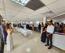 Imagem que mostra a inauguração da sala da DPE-PR no Fórum de Cambé, onde a defensora pública Aline Bastos discursa para várias pessoas que assistem a cerimônia. Todos estão em pé, em um hall amplo e bem iluminado, dentro do Fórum. Há também duas mesas no centro, com comidas e bebidas.