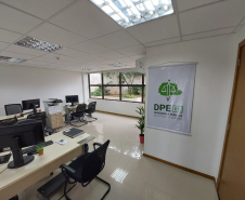 Imagem que mostra outra sala da DPE-PR no Fórum de Cambé, onde há um banner com o logo da Defensoria na parede ao lado da porta e três mesas com computadores e cadeiras para pessoas serem atendidas. Ao fundo é possível ver uma grande janela que mostra uma parte externa do Fórum, onde há algumas plantas em um canteiro.