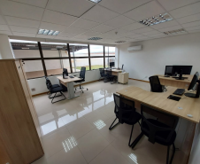 Imagem que mostra outra sala da DPE-PR no Fórum de Cambé, onde há um armário e quatro mesas com computadores e cadeiras para pessoas serem atendidas. Ao fundo é possível ver uma grande janela que mostra uma parte externa do Fórum.