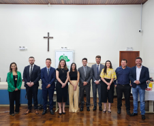 Imagem que mostra representantes da Defensoria e autoridades convidadas na inauguração da sala da DPE-PR em Jandaia. As pessoas, 4 mulheres e 6 homens, estão lado a lado e sorriem. Elas estão em uma sala ampla do Fórum, onde foi realizada a solenidade. Atrás delas é possível ver um banner com o logo da Defensoria e um crucifixo. 