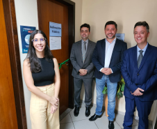 Imagem que mostra a defensora pública Maria Luiza Valverde, o defensor público-geral, André Giamberardino, e o prefeito de Jandaia do Sul, Lauro Junior, diante da porta da sala da Defensoria no Fórum de Jandaia. Na porta, há uma placa que diz “Defensoria Pública” e cruzando a porta de um lado a outro há uma fita de cetim de cor verde, com um laço ao meio. Todos sorriem.