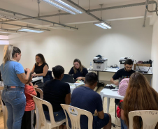 Imagem com a foto de usuários e usuárias da Defensoria recebendo o atendimento sentamos em cadeiras e mesas brancas dentro de uma sala. 