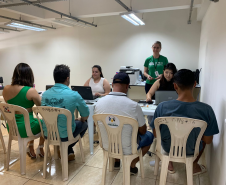 Imagem com a foto de usuários e usuárias da Defensoria recebendo o atendimento sentamos em cadeiras e mesas brancas dentro de uma sala. 