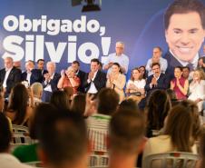 Imagem colorida horizontal. No primeiro plano se vê o público observando autoridades no lançamento do projeto de construção do Centro de Reabilitação Silvio Santos. Ao fundo cartaz com a imagem do apresentador onde se lê: Obrigado, Silvio.