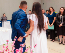 Imagem que mostra um dos casais, de costas. Ele usa um terno azul e ela um vestido branco. Ela está com o cabelo solto, liso, que vai até o meio das costas. Ao fundo, as autoridades presentes assistem a cena e sorriem. Arranjos com flores rosas em arranjos compõem a cena.