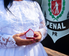 Imagem que mostra uma noiva com um vestido branco com aplicações de renda. Ela segura uma pequena caixa vermelha com duas alianças. Ao fundo é possível ver o brasão do DEPEN em uma parede.