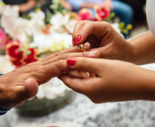 Imagem que mostra uma das noivas colocando a aliança no dedo do noivo. O fundo mostra flores desfocadas. É possível ver parte do vestido dela, branco. Ela tem as unhas pintadas com esmalte vermelho.