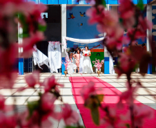 Imagem que mostra as noivas em fila preparadas para a entrada. O ambiente está decorado com panos brancos, flores rosas e um tapete vermelho.