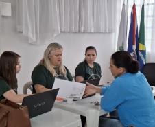 Imagem que mostra um dos atendimentos do evento. Três pessoas da Defensoria atendem uma mulher. Uma delas entrega um documento para a mulher que está sendo atendida.