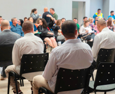 Imagem que mostra os noivos sentados em cadeiras pretas, de costas. Ao fundo é possível ver as autoridades presentes.