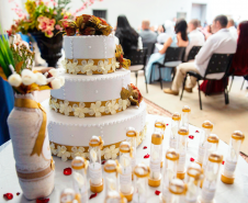 Imagem que mostra uma mesa decorada com um bolo, arranjo de flores e lembrancinhas em tons de branco e dourado. Ao fundo é possível ver os casais sentados, de costas.