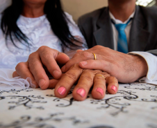 casal com as mãos sobre a mesa mostrando as alianças. eles usam trajes de terno e gravata e vestido de noiva.