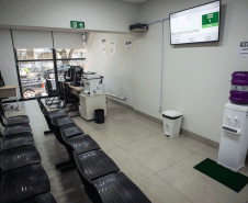 Espaço de espera com fileiras de cadeiras pretas para o público, um bebedouro de água, uma lixeira e um painel eletrônico na parede exibindo senhas de atendimento; no fundo, uma grande janela de vidro permite a visão da rua.