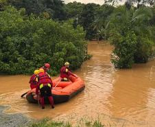 Imagem horizontal colorida em que se vê bombeiros em um bote inflável entrando em área alagada pelas enchentes no Litoral do Paraná. 
