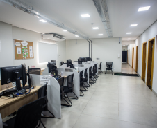 Sala ampla e bem iluminada com várias mesas de atendimento organizadas em fileiras, cada uma equipada com computador e cadeiras para os atendentes e assistidos; ao fundo, um corredor leva a outras salas.