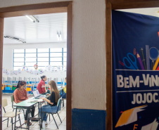 Imagem que mostra o interior de uma sala de aula vista através de uma porta entreaberta. Na sala, há pessoas sendo atendidas por agentes da DPE-PR. A sala possui janelas grandes com grades, permitindo a entrada de luz natural. Há decorações coloridas penduradas nas janelas e nas paredes. À direita, em primeiro plano, parte de uma faixa azul escura está visível, pendurada na porta, com a inscrição "BEM-VINDOS JOJOCA" em letras brancas grandes, acompanhada de desenhos coloridos de materiais escolares.