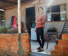 Imagem que mostra a senhora Amélia sorrindo em frente de sua casa. Ela tem cabelos escuros e veste uma blusa de manga comprida cor de telha, calças pretas e chinelos escuros. Ela está em uma área externa coberta, com piso de ladrilho claro. Ao fundo, à esquerda, há uma área mais aberta com vegetação e roupas penduradas em um varal improvisado. A casa é simples, tem paredes rebocadas e uma janela gradeada com cortinas brancas. A luz do sol entra pela lateral direita da imagem, criando um efeito de brilho.