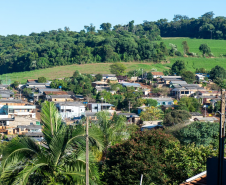 Imagem que mostra uma vista panorâmica do bairro São João, em Pato Branco. As casas são simples, com telhados variados em tons de cinza e marrom, e estão dispersas entre a vegetação exuberante, incluindo árvores altas e palmeiras em primeiro plano. As colinas ao fundo são cobertas por uma mistura de floresta densa e áreas de pastagem mais claras. A luz do sol incide sobre a cena, realçando as cores da natureza e as construções.