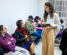 Imagem colorida horizontal em que se vê servidora da DPE-PR entregando as senhas e fazendo a orientação dos assistidos. 