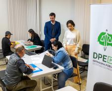 Imagem colorida horizontal em que se vê o banner da DPE-PR em primeiro plano e ao fundo o DPG acompanhando os atendimentos do mutirão.