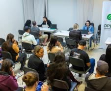 Imagem colorida horizontal em que se vê uma sala cheia de pessoas e ao fundo a equipe da DPE-PR realizando atendimento. 
