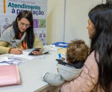 Imagem colorida horizontal em que se vê uma moça de costas segurando um bebê também de costas e ao fundo servidora do MPPR realizando atendimento durante o mutirão conjunto com a DPE-PR. 