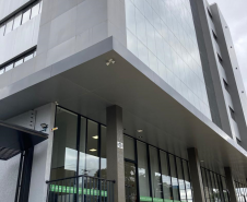 Imagem que mostra a nova sede da Defensoria em Foz do Iguaçu, com uma perspectiva mais ampla da fachada do prédio. O edifício apresenta um design moderno com múltiplas camadas e uma combinação de grandes painéis de vidro espelhados. Uma faixa horizontal verde clara com o logo da DPE-PR percorre a fachada. Uma rampa acessível com piso tátil amarelo leva à entrada principal e há também um corrimão de metal preto. O céu está nublado, o que confere uma iluminação difusa ao ambiente externo. 