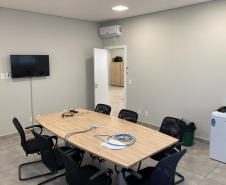 Imagem que mostra uma sala de reunião da nova sede da Defensoria em Foz do Iguaçu. Ela está equipada com uma mesa grande de madeira clara no centro. Ao redor da mesa, há várias cadeiras pretas e em cima da mesa, alguns cabos azuis de conexão à internet e documentos estão visíveis. Em uma das paredes, há uma televisão de tela plana montada e, à direita da imagem, um frigobar branco.