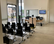 Imagem que mostra a área de recepção da nova sede da Defensoria em Foz do Iguaçu. À esquerda, grandes portas de vidro permitem a entrada de luz natural e oferecem vista para o exterior, onde é possível ver uma rua e vegetação. O ambiente possui fileiras de cadeiras pretas com estrutura metálica, voltadas para uma área de atendimento. Ao fundo, há mesas de madeira clara com computadores e monitores pretos, indicando estações de trabalho para atendimento ao público. 
