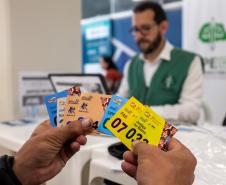 Em primeiro plano se vê as mãos de um assistido segurando as senhas de diversos serviços oferecidos no Registre-se e ao fundo desfocado o defensor público que fazia o seu atendimento. 