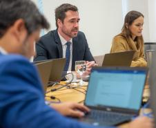 A imagem mostra pessoa de costas em primeiro plano e ao fundo se vê um homem e uma mulher; todos estão usando laptops.