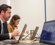 Um homem de terno e uma mulher acompanham reunião ao computador e em primeiro plano tela de laptop em que aparecem os participantes online da reunião. 