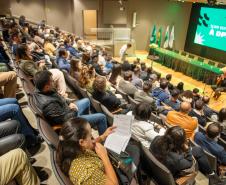 Imagem mostra o auditório quase cheio para a posse dos servidores, no palco ocorre uma apresentação músicas de violão e voz. Ao fundo mesa para as autoridades e o telão com logo da DPE-PR. 