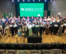 Grupo de Servidores empossados posam juntos para foto no palco de auditório. Ao fundo tela de Led mostra a logo da instituição.  
