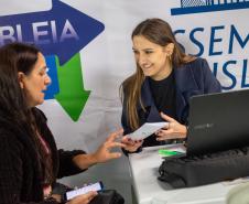 Imagem mostra duas mulheres sentadas, uma servidora ao computador entrega documentos a uma assistida.  