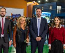 Quatro pessoas posam para foto, dois homens de terno e duas mulheres. Ao fundo se vê grande número de pessoas circulando pelo evento. 