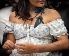 Imagem em detalhe de uma mulher indígena onde se vê o vestido, os braços com pintura corporal e brinco de penas. 
