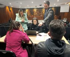 De costas um casal sentado recebe orientações da equipe da DPE-PR, formada por duas mulheres e um homem. Ele aponta a tela de um computador. 