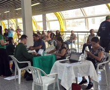 Imagem mostra um grande grupo de pessoas, algumas em pé, outras sentadas à mesa, em mutirão da Defensoria realizado em terminal de ônibus.