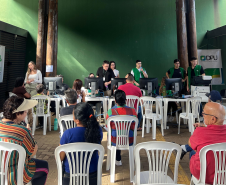 Imagem mostra grupo de pessoas sentadas em cadeiras de plástico no primeiro plano e ao fundo se vê pessoas sendo atendidas em mesas por defensores e servidores da DPE-PR. 