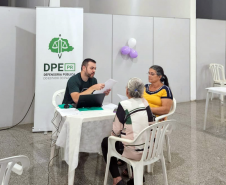 Imagem mostra duas mulheres, um idosa, sentadas à mesa conversando com homem jovem usando o computador, Ao fundo se vê banner da Defensoria com a logo da instituição.