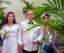 Casal desfocado caminha em tapete vermelho em cerimônia de casamento. No primeiro plano aparece a folhagem da decoração.  