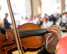 Violino sendo tocado em close-up. Ao fundo aparecem os casais e familiares em desfoque. 