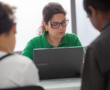 Casal de costas sentado em primeiro plano e ao fundo servidora da DPE-PR usando camisa verde e óculos realiza atendimento ao computador.  