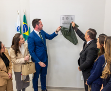 A imagem captura o momento do descerramento da placa inaugural. O Defensor Público-Geral, à esquerda, e o vice-prefeito de Foz do Iguaçu, à direita, seguram juntos um tecido verde-escuro, revelando a placa fixada na parede. Ao redor deles, um grupo de convidados, majoritariamente mulheres, observa a cena com sorrisos e expressões de satisfação.